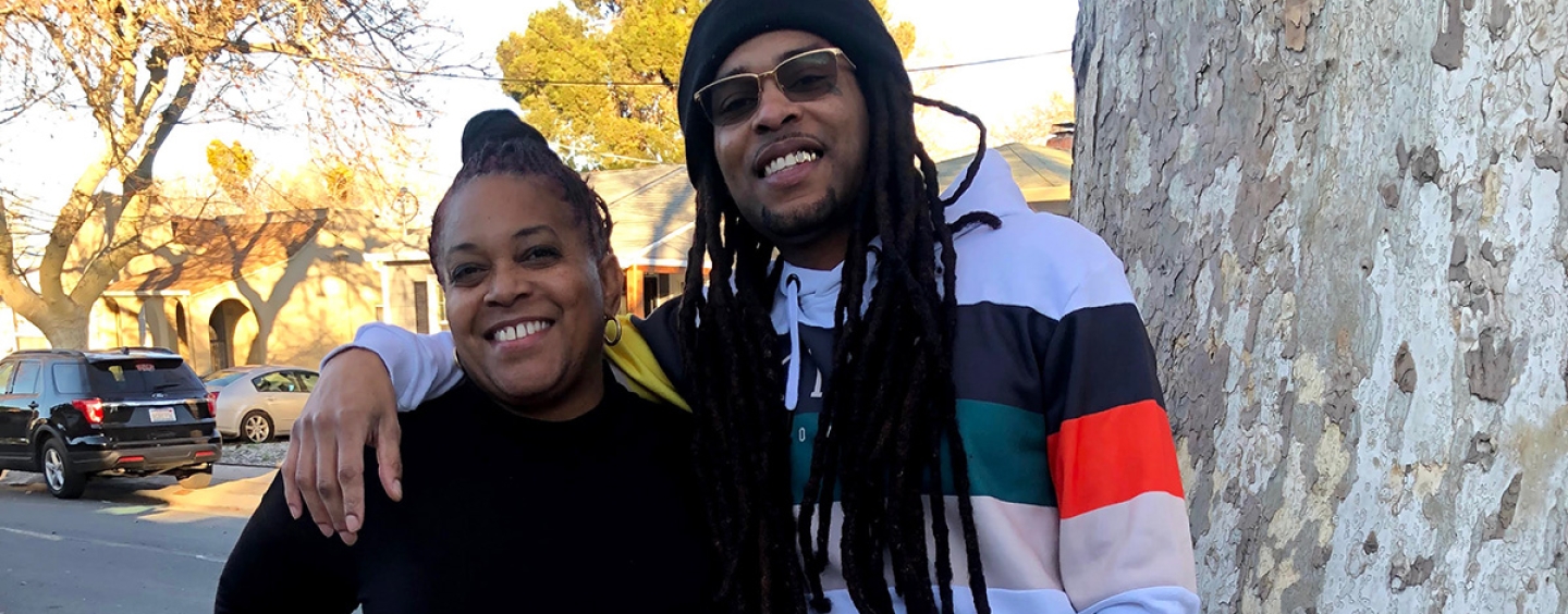 Marcus Angelo Bryant stands to the right of his mother, Theresa Thompson, with his right arm around her shoulders while both smile and stand next to a tree on a sunny day