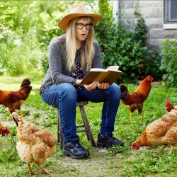 Nona Caspers reads from a book, sitting on the lawn of an old house, surrounded by chickens